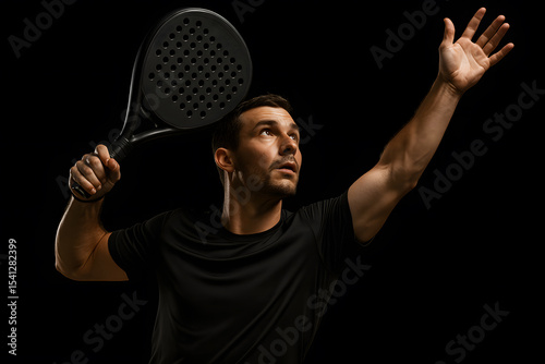 A young adult male, appearing focused and athletic, holds a padel racket overhead, ready to serve, against a black background