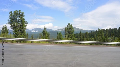 POV driving through forested mountain road on sunny summer day. View from inside moving car, with green trees, clear blue sky, and distant snow-capped peaks. Scenic peaceful travel footage in Siberian