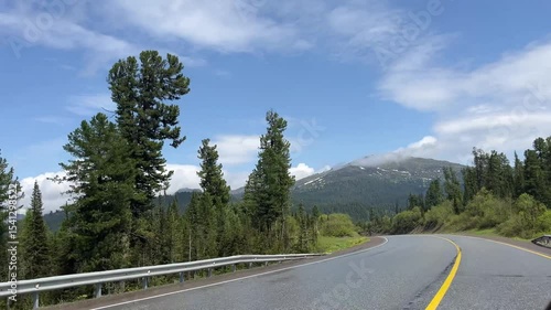 POV driving through forested mountain road on sunny summer day. View from inside moving car, with green trees, clear blue sky, and distant snow-capped peaks. Scenic peaceful travel footage in Siberian