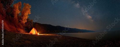 A glowing tent on a lakeshore under a star-filled night sky with the Milky Way visible, surrounded by autumn trees and distant hills.