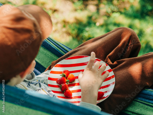 Young Boy in Cap Eating Strawberry in Hammock