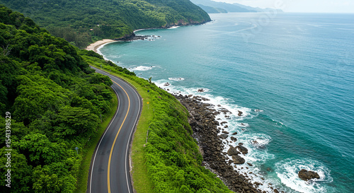 Aerial view of a scenic winding asphalt road curving smoothly along a lush green coastal cliffside, bordered by thick tropical vegetation on one side and a calm