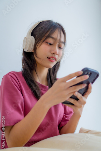 Adorable Asian little girl wearing headphones sitting on bed in her bedroom, happily listening to music and holding a smartphone. Cute child relaxing and enjoying playful, joyful moment at home.