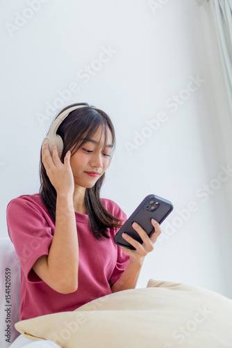 Adorable Asian little girl wearing headphones sitting on bed in her bedroom, happily listening to music and holding a smartphone. Cute child relaxing and enjoying playful, joyful moment at home.
