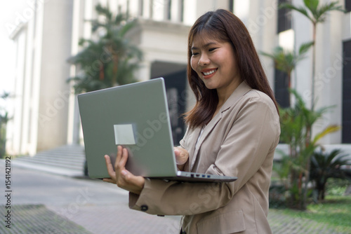 Young happy Asian businesswoman working on laptop in city. Confident female executive using computer while standing outdoors in urban business district, multitasking and managing work on the go.