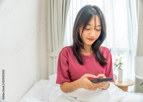 Asian young girl using smartphone texting and chatting with friend in bedroom at home. Relaxing woman wearing pink shirt sit on the bed using mobile phone to communicate in the morning at house.