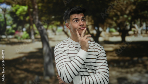Young man with thoughtful expression in a striped shirt contemplating in a sunny park setting surrounded by trees showcasing a serene outdoor environment.