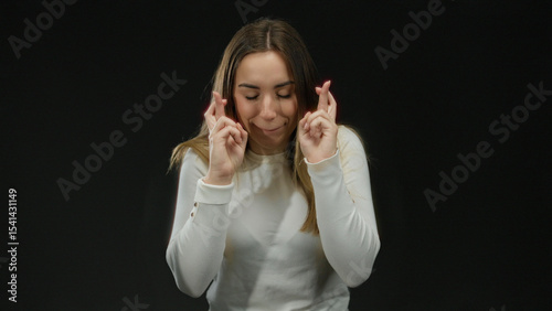 Young woman with blonde hair crossing fingers hopefully against isolated black background in candid portraits.