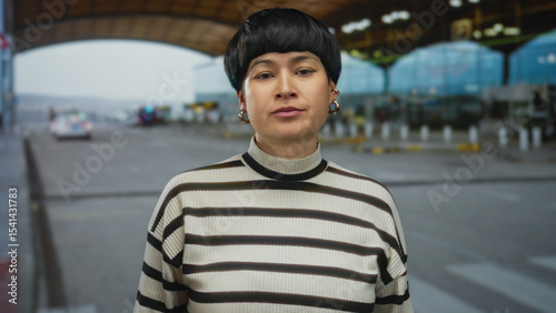 Woman gesturing at an airport outdoor setting, expressing displeasure with crossed arms and stern expression, wearing striped sweater in a modern open-air terminal.