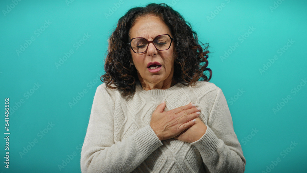 Woman experiencing discomfort holding chest with curly hair against blue background wearing sweater looking distressed isolated mature hispanic adult Woman experiencing discomfort holding chest with curly hair against blue background wearing sweater looking distressed isolated mature hispanic adult