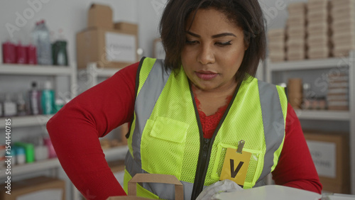 Woman wearing reflective vest and gloves prepares food packages in indoor charity setting with cardboard boxes and donations visible behind her.