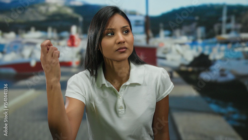 Woman gesturing at seaside port with boats in background under sunny sky, expressing emotions in outdoor setting, wearing white shirt, young and hispanic.