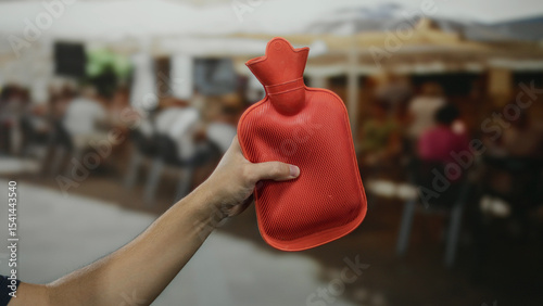 Hand of a man holding a red hot water bottle on a busy outdoor restaurant terrace in the city.