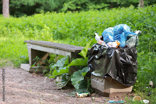 An Overflowing Public Trash Can With Plastic Bags And Litter Spilling Out, Next To A Wooden Bench In A Park.