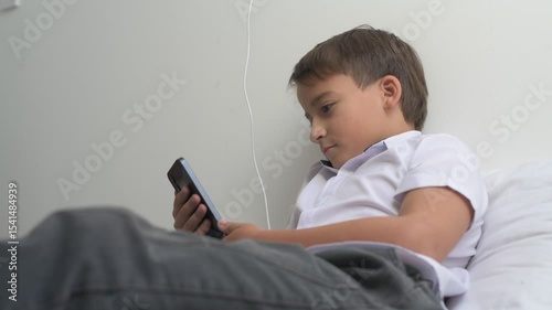 Little schoolboy is engrossed in watching the screen of mobile phone while lying on bed at home