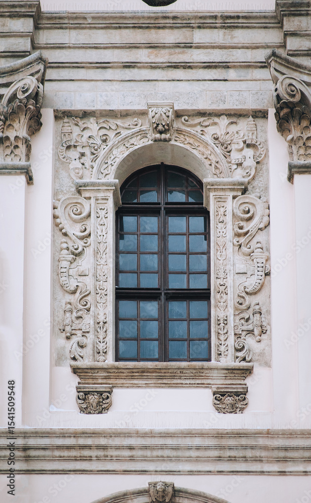 Antique baroque window with a wooden brown frame, decorated with decorative stucco on a gray wall. Jesuit Church in Lvov. Antique baroque window with a wooden brown frame, decorated with decorative stucco on a gray wall. Jesuit Church in Lvov.
