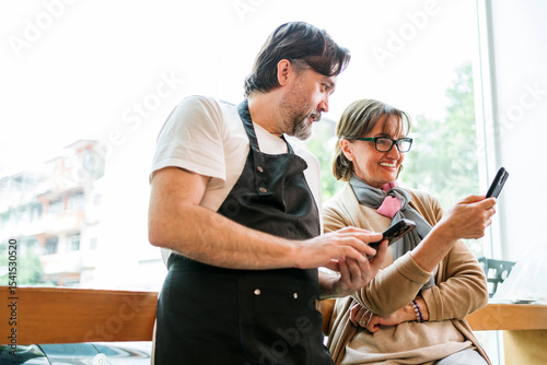 Cafe owner and business advisor discussing marketing or finance using smart phones. Both are smiling and engaged, symbolizing teamwork, strategy, and modern small business collaboration.