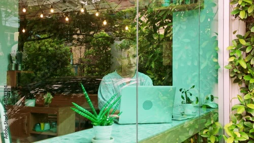 Thoughtful cafe owner works on a laptop while gazing out the window in a lush, plant-filled shop. Scene captures entrepreneurship, creativity, and peaceful productivity in a cozy urban coffee space.