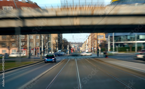 Motion blur effect captures a blue car driving on urban tram tracks under a bridge, with surrounding city buildings and light traffic. Dynamic street scene showing movement and speed in a metropolitan