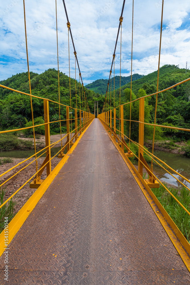 Obraz premium Yellow Iron Pedestrian Bridge Across Mountain River Against Mountain Backdrop In Vietnam