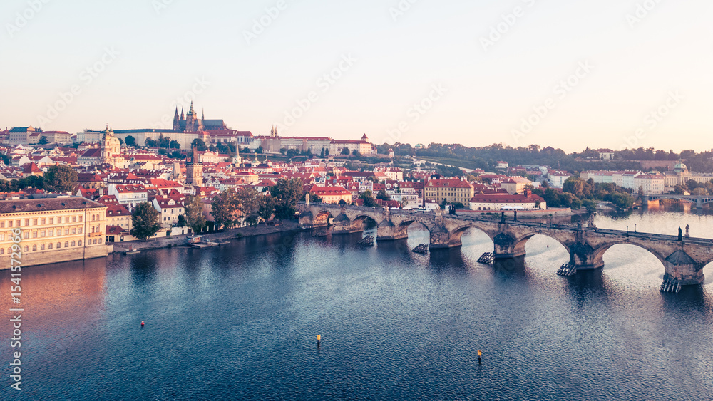 Fototapeta premium As dawn breaks over Prague, the golden hues illuminate the medieval architecture along the Vltava River, with the iconic Charles Bridge and Hradcany Castle in view, creating a breathtaking panorama.