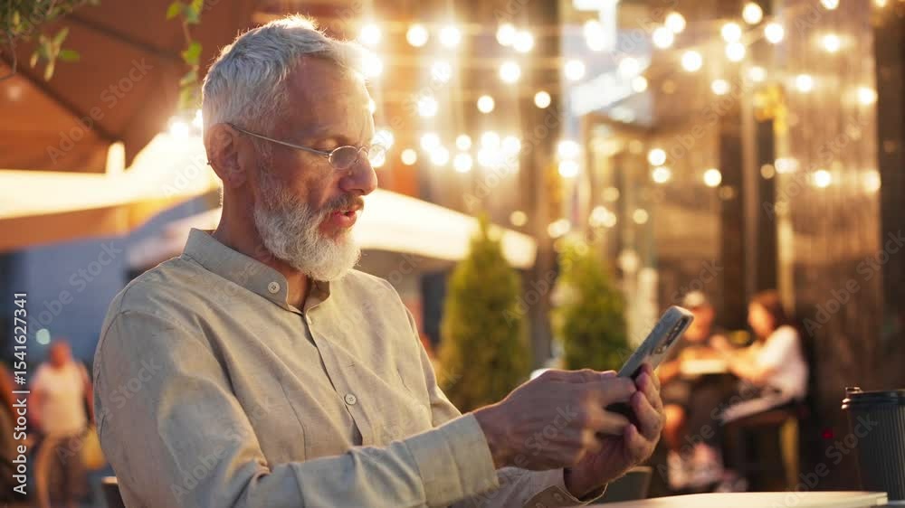 Expressive Caucasian man with white beard and glasses reading something on smartphone display. Outdoor cafe. Saying text aloud while gesturing enthusiastically under warm string lights in evening.