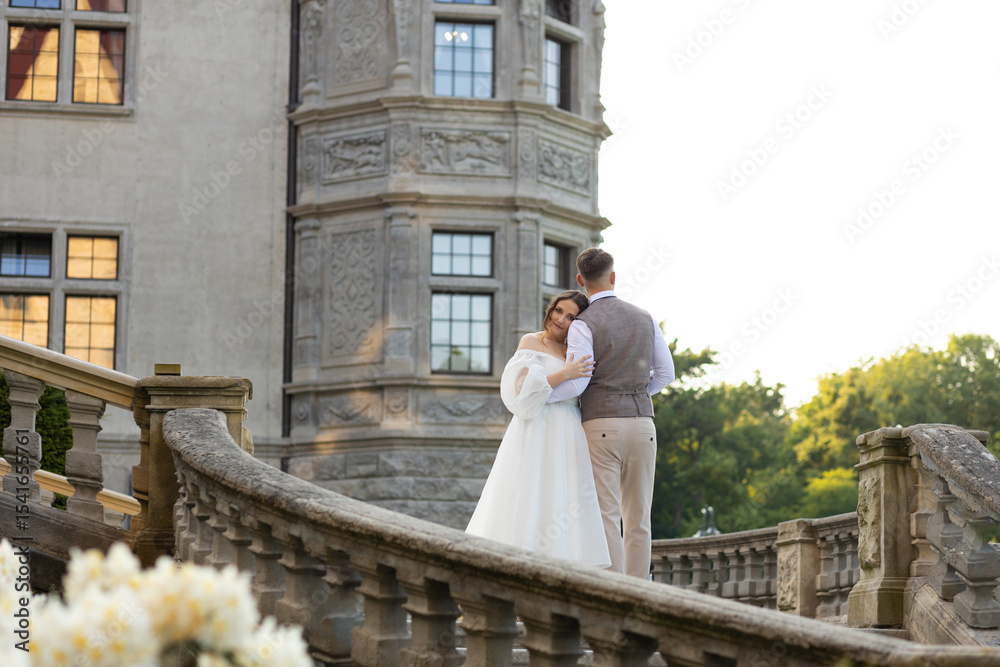 Obraz premium Stylish exterior of a historic castle with newlyweds standing on the steps. Bride resting her head on her husband's shoulder against the background of the castle.