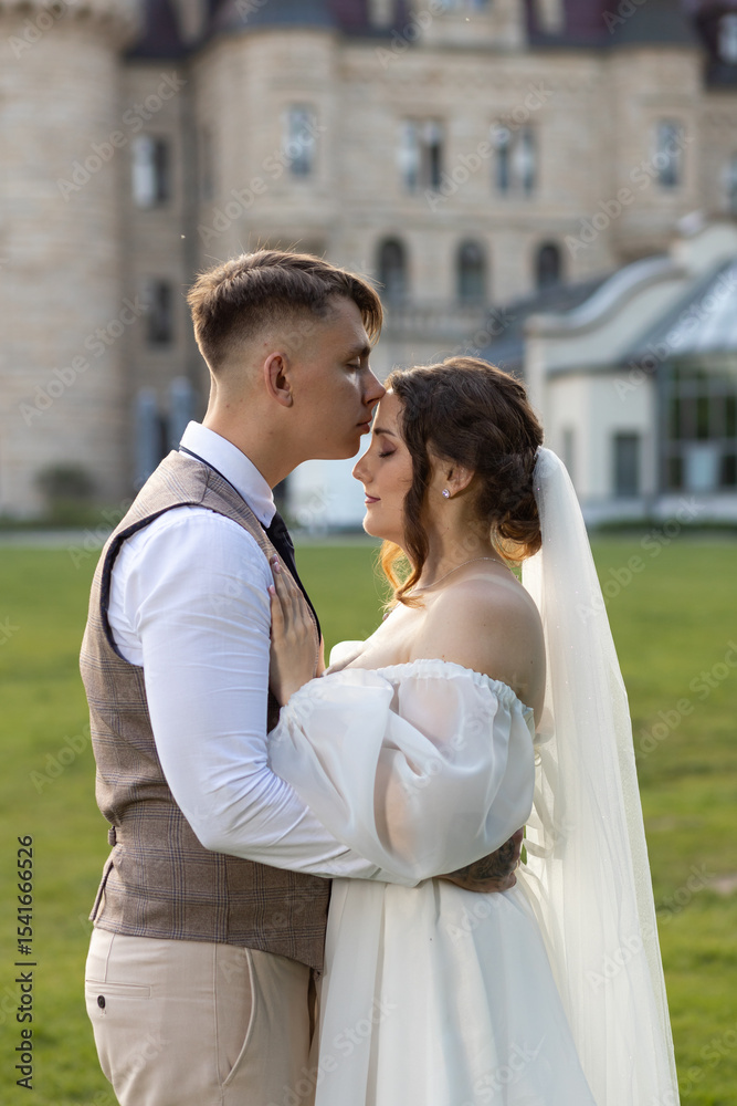 Naklejka premium The groom kisses the bride on the forehead and hugs her waist. An incredibly tender wedding photo against the backdrop of the Moszna Castle. A harmonious couple of newlyweds.