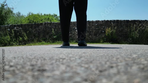 Stock video: A ground level view of an unidentified male walking slowly walking across a road, away from the camera, towards an old bridge wall.