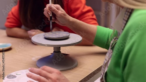 Skilled pottery instructor guiding student while hand painting delicate ceramic plate on rotating support in creative ceramic studio workshop