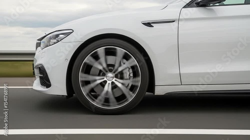 Close Up of White Sedan Car in Motion on Asphalt Road Under Sky with Blurry Background and Rotating Wheels