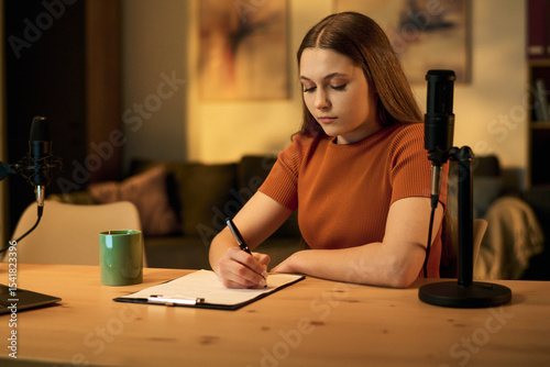 Caucasian teenage girl sitting at desk writing notes on clipboard preparing for podcast recording with professional microphone