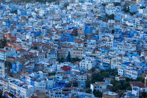 An expansive aerial view captures the dense, picturesque blue-washed buildings of Chefchaouen, Morocco, sprawling across a hillside under a soft, diffused light.