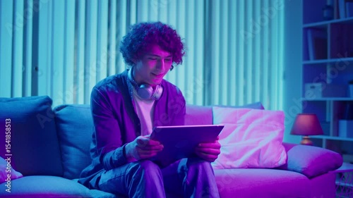 Mental Health, Digital Connection, Emotional Release. Young Man Sits on Sofa at Night, Laughing at Tablet Screen. Moment of Joy, Relaxation, and Connection in Digital Daily Life