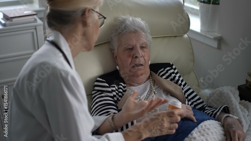 A mature woman in a comfortable home chair listens as a caregiver explains medication instructions during a house visit.