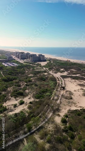 Aerial view of a modern beachside development with apartment buildings, sand dunes, and a wide sandy beach under a clear blue sky. There are people walking, cyclists and cars driving along the road.