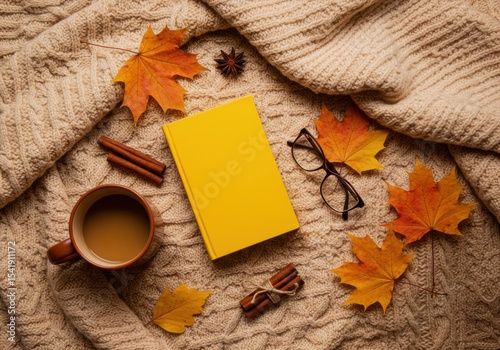 A cozy autumn scene featuring a yellow book, a cup of coffee, glasses, cinnamon sticks, and colorful maple leaves on a knitted blanket.