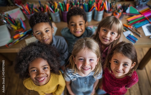 Five diverse children smile up at the camera, surrounded by colorful art supplies on a wooden table, showcasing creativity and joy in a classroom setting.