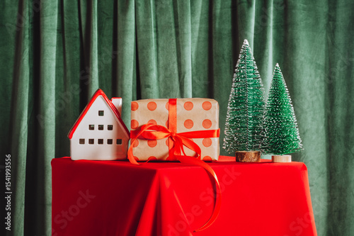Wallpaper Mural Christmas composition with gift box, ceramic house, and green mini trees on red tablecloth. Cozy festive home decor Torontodigital.ca