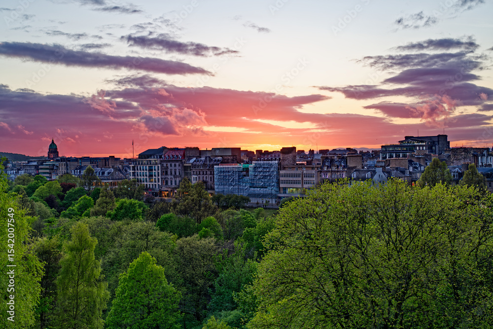 Fototapeta premium Beautiful sunset with orange pink sky above buildings on Princess street in Edinburgh, UK. Colorful sky with clouds. Above public park with green trees. Edinburgh, UK, 05.04.2025