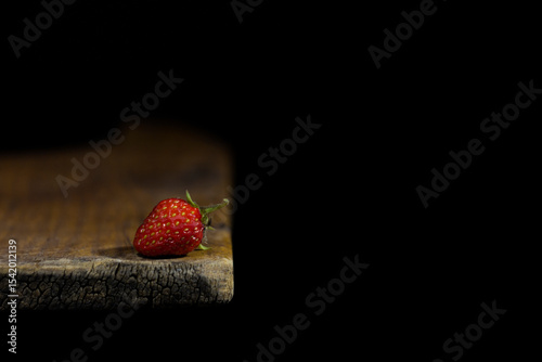 The image shows strawberries lying on the edge of a wooden surface. The background is dark, which emphasizes the brightness and texture of the berry.