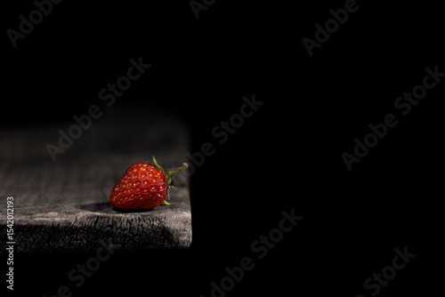 The image shows a single strawberry lying on the edge of a wooden surface. The background is black, which creates a contrast with the bright red berry illuminated by the light.