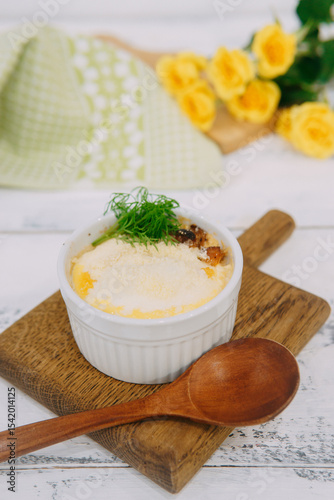 Baked casserole served in a white ramekin garnished with fresh green dill on a wooden board, accompanied by yellow roses and soft light.