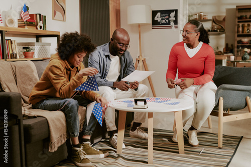 Papier peint Teenage boy sitting on sofa assembling American flags with father and mother smi