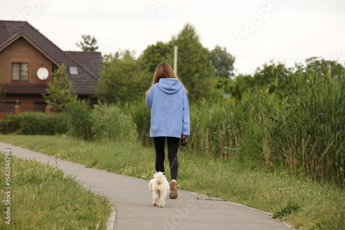 A young girl in a blue jacket and black pants walks home with her dog along an alley among the greenery. Returning from a walk with a pet. Animals care.