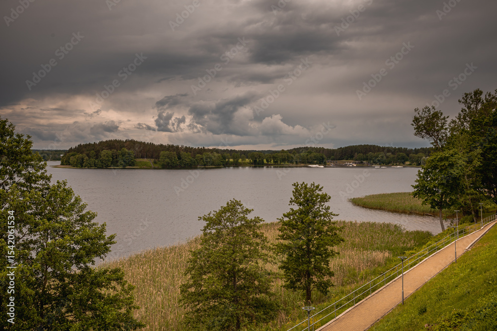 Fototapeta premium A beautiful lake in a forested area with aquatic plants, canes, reeds and trees on the shore under cloudy stormy sky