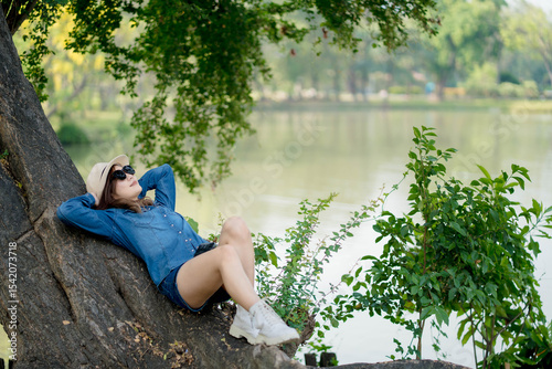Young woman relaxing on a tree trunk by the lake in a peaceful natural park on a sunny day, wearing a hat, sunglasses, and denim outfit photo