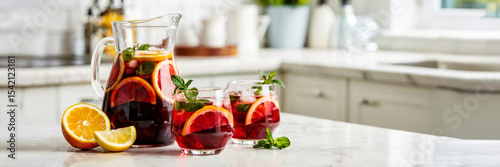 A glass pitcher of red sangria with citrus slices and mint, placed next to two glasses on a white marble kitchen countertop, long banner