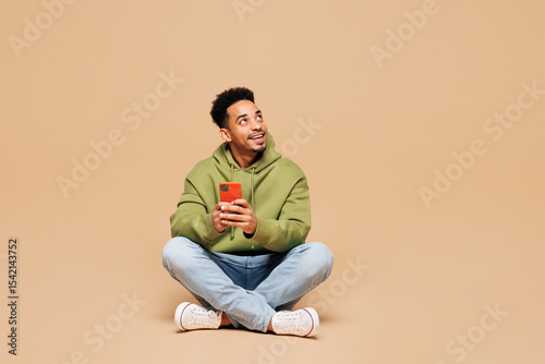 Full body young man of African American ethnicity wears green hoodie casual clothes sit hold in hand use mobile cell phone look aside isolated on plain pastel light beige background Lifestyle concept