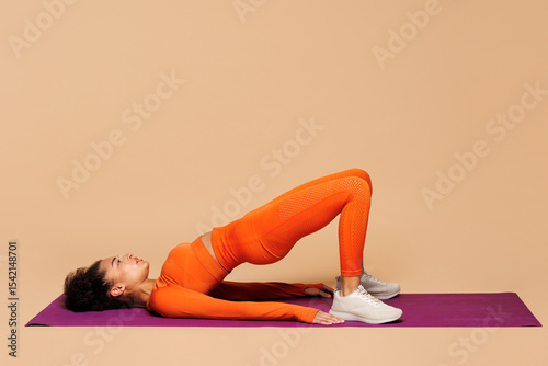 Full body young fitness trainer instructor sporty woman wear orange clothes spend time in gym do glute bridge exercise on yoga mat o isolated on plain beige background. Workout sport fit abs concept.